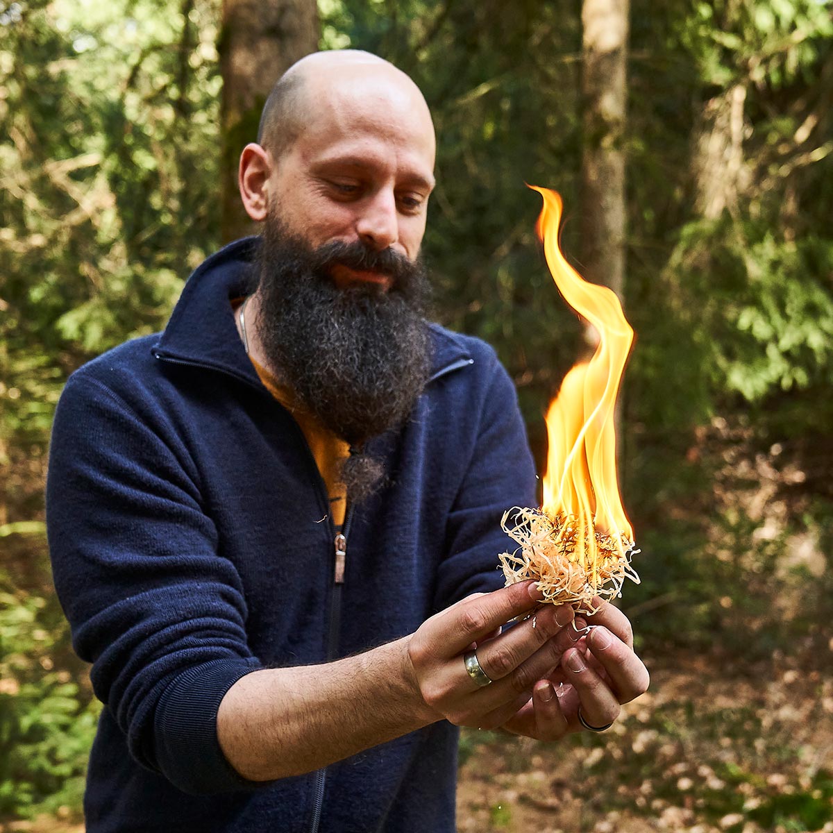 Transianer Richu steht im Wald und hält kleines Zündmaterial in der Hand 