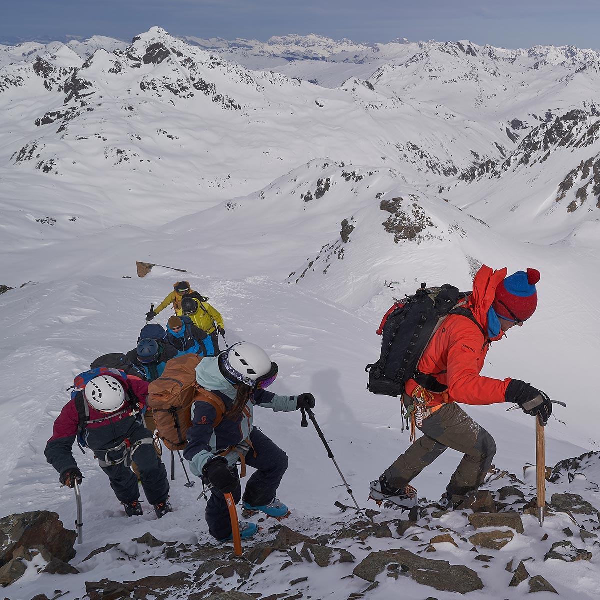 Eine Gruppe Skitourengeher:innen mit Bickel und Steigeisen auf den letzten Metern zu Gipfel, im Hintergrund verschneite Berge