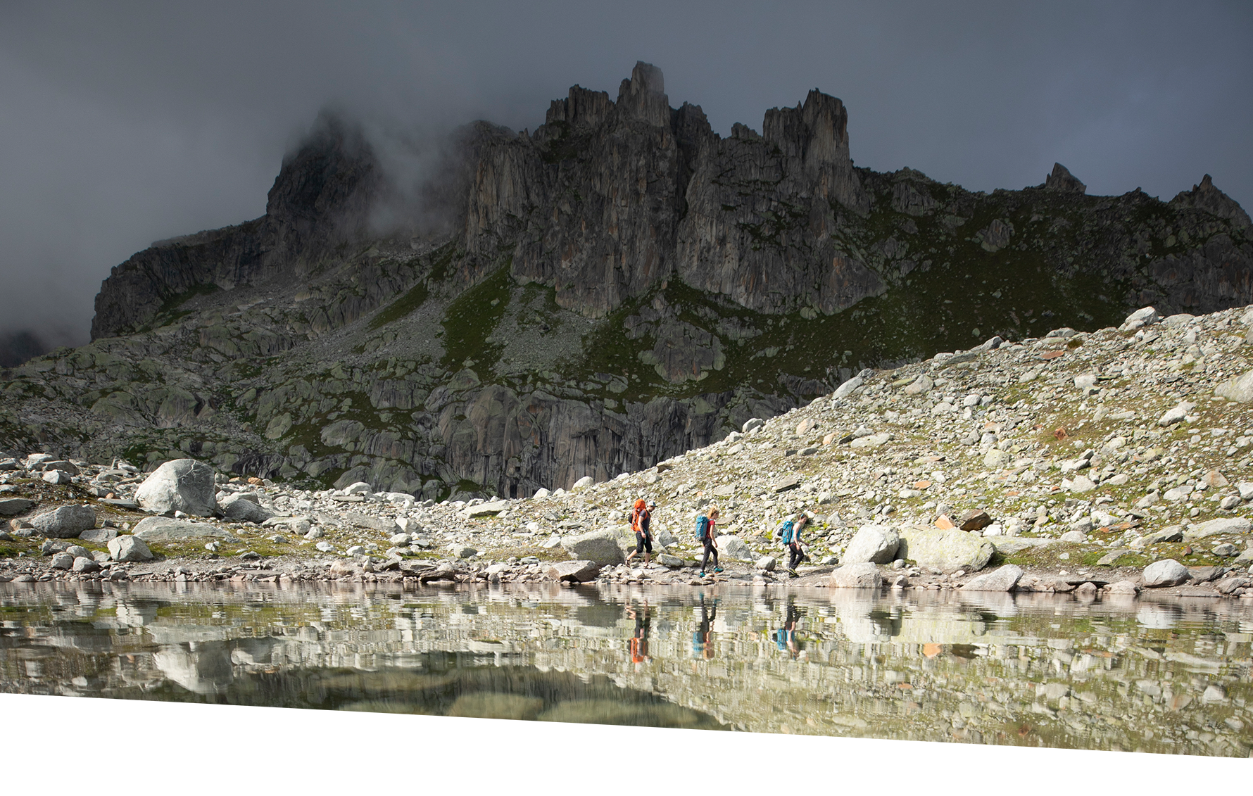 Drei Personen wandern an einem Bergsee entlang, im Hintergrund eine mächtige Felswand 