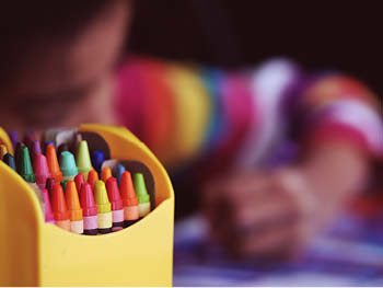 Closeup of a box of crayons of assorted colors, with a child in the background