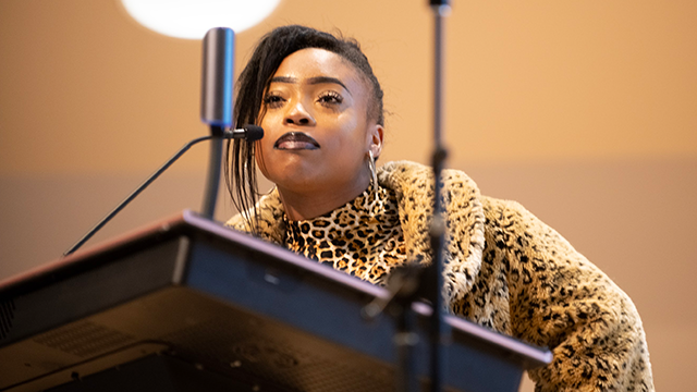 A speaker at a podium, wearing leopard print and with dark hair sloped to on side of their face.