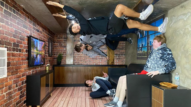 A room with a TV and a student sitting on a couch; other students appear to be standing at weird angles with their feet on the wall or climbing on the ceiling.