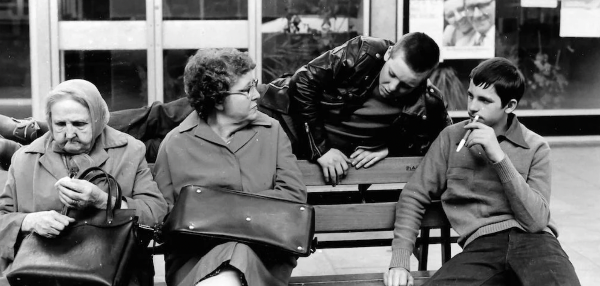 A black and white photo of four light skinned people on a bench, 3 looking at eachother and an elderly woman looking away