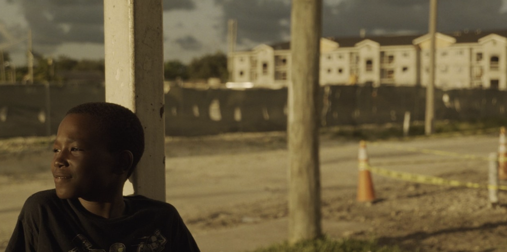 : a dark-skinned boy looks out over the street, traffic cones and construction in the background