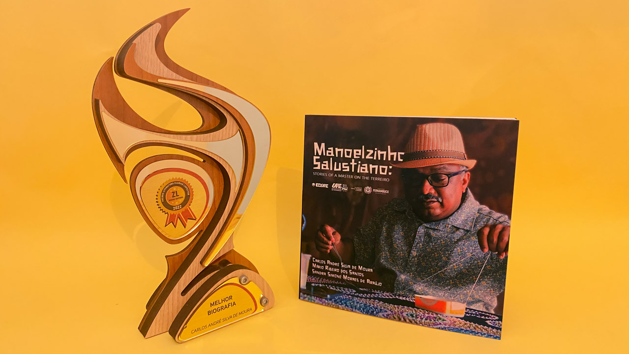 Foto do troféu do Prêmio Literatura Clarice Lispector e da capa do livro “Manoelzinho Salustiano: histórias de um mestre no terreiro”