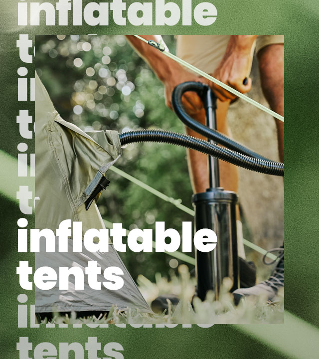 Close-up of a camping scene showing a green inflatable tent being filled with air using a black hand pump. A person’s lower legs and hands are visible while operating the pump, with the hose connected to the tent’s air valve. The setting appears outdoors among trees, with soft, natural light and blurred greenery in the background. 