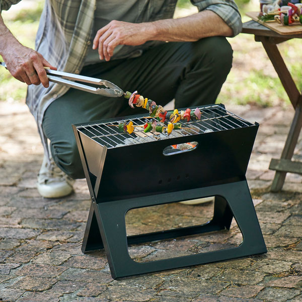 A compact black portable grill sits on a paved surface, with skewers of vegetables cooking over the grate. A person uses tongs to turn the skewers, while a small side table with food appears in the background.
