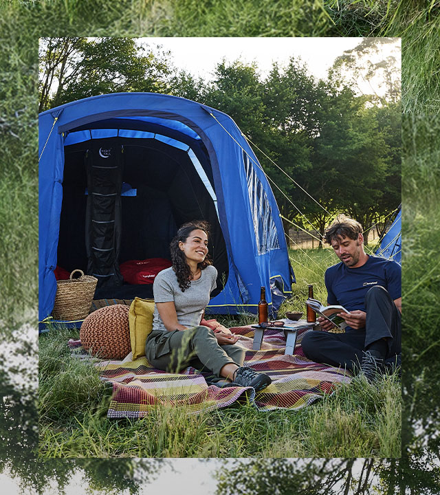 Two people sit on a picnic blanket in a grassy campsite beside blue tents, with trees in the background and camping gear arranged nearby.
