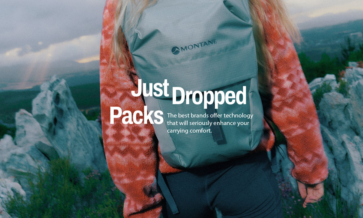 Close-up view of a person from behind wearing a light grey Montane backpack and an orange patterned fleece jacket, walking outdoors among rocky terrain and greenery, with blurred mountains in the background. 