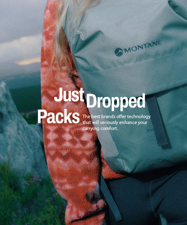 Close-up view of a person from behind wearing a light grey Montane backpack and an orange patterned fleece jacket, walking outdoors among rocky terrain and greenery, with blurred mountains in the background. 