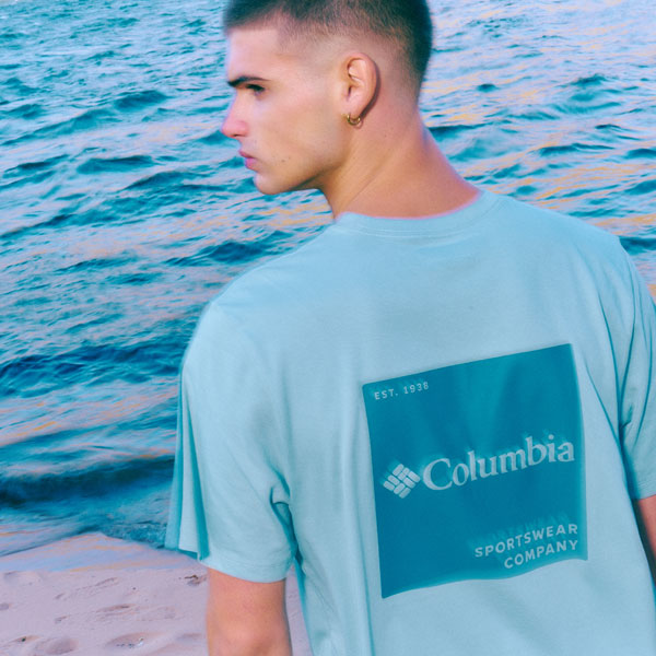 A photo of a man posing by the sea wearing a short sleeve t-shirt.