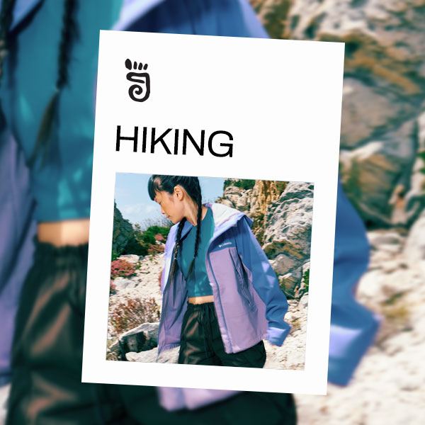 A woman hiking on a sunny trail wearing a purple jacket and shorts.