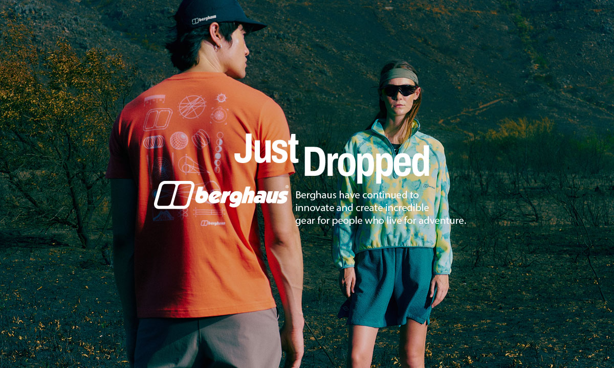 Two people stand facing each other in an outdoor, rugged landscape with dark terrain and sparse vegetation. The person in the foreground wears an orange T-shirt with a graphic design and the Berghaus logo on the back, along with a black cap. The second person, positioned further back, wears a patterned green and yellow jacket, dark shorts, sunglasses, and a headband. 