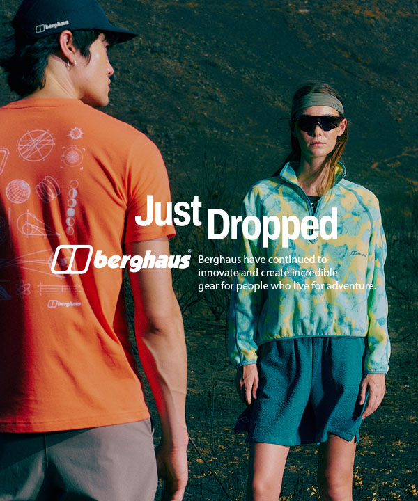 Two people stand facing each other in an outdoor, rugged landscape with dark terrain and sparse vegetation. The person in the foreground wears an orange T-shirt with a graphic design and the Berghaus logo on the back, along with a black cap. The second person, positioned further back, wears a patterned green and yellow jacket, dark shorts, sunglasses, and a headband. 