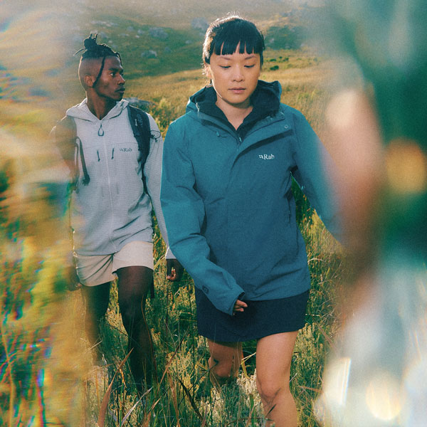 Two hikers wearing lightweight outdoor jackets walking through grassy hillside terrain with mountains in the background.