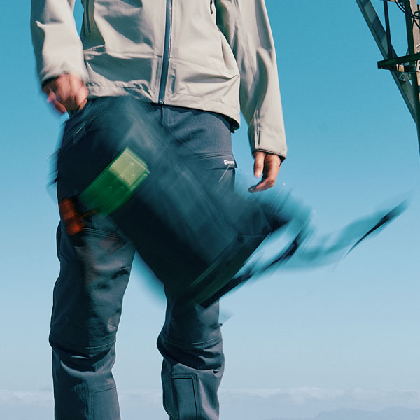Person holding a backpack while standing outdoors, wearing technical hiking trousers against a clear blue sky.”