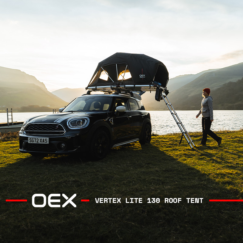 Black Mini Countryman with an OEX Vertex Lite 130 rooftop tent parked beside a lake at sunset, with mountains in the background and a person walking near the ladder.