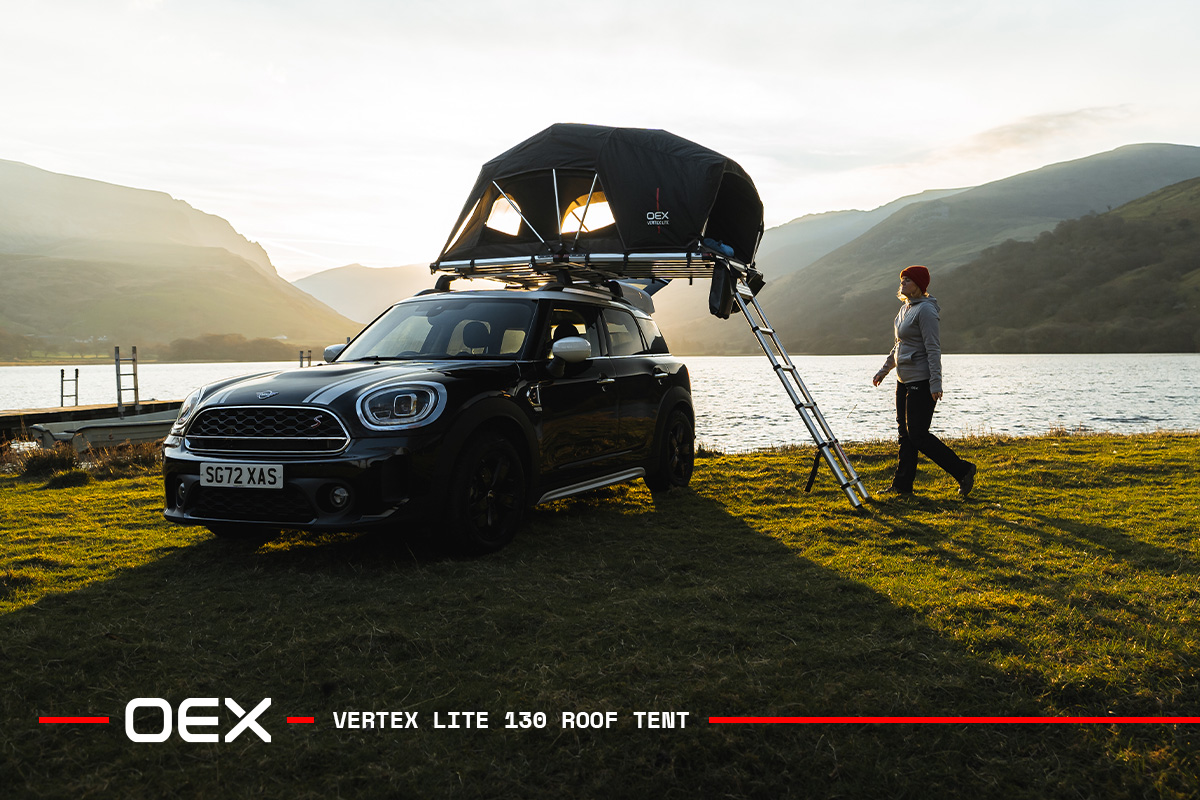 Black Mini Countryman with an OEX Vertex Lite 130 rooftop tent parked beside a lake at sunset, with mountains in the background and a person walking near the ladder.