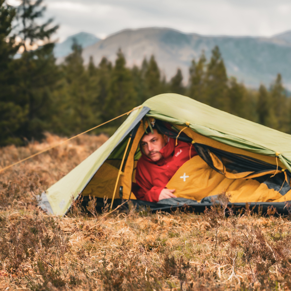 Person lying inside a small green tent on grassy terrain, wrapped in an orange sleeping bag, with trees and mountains in the background.