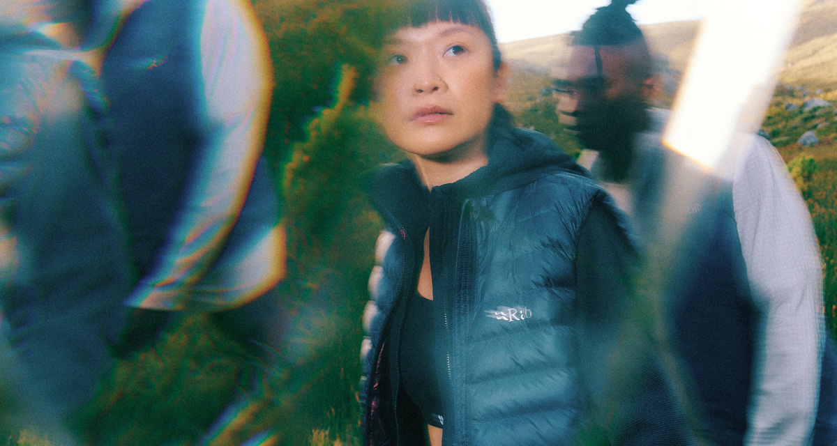 Person wearing a dark puffer vest standing outdoors among blurred figures, with a stylised lens effect and a natural landscape in the background.