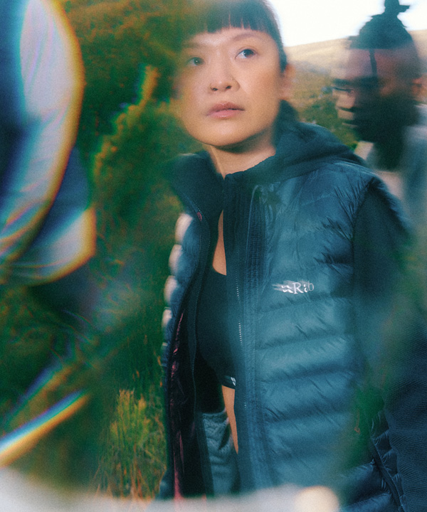 Person wearing a dark puffer vest standing outdoors among blurred figures, with a stylised lens effect and a natural landscape in the background.