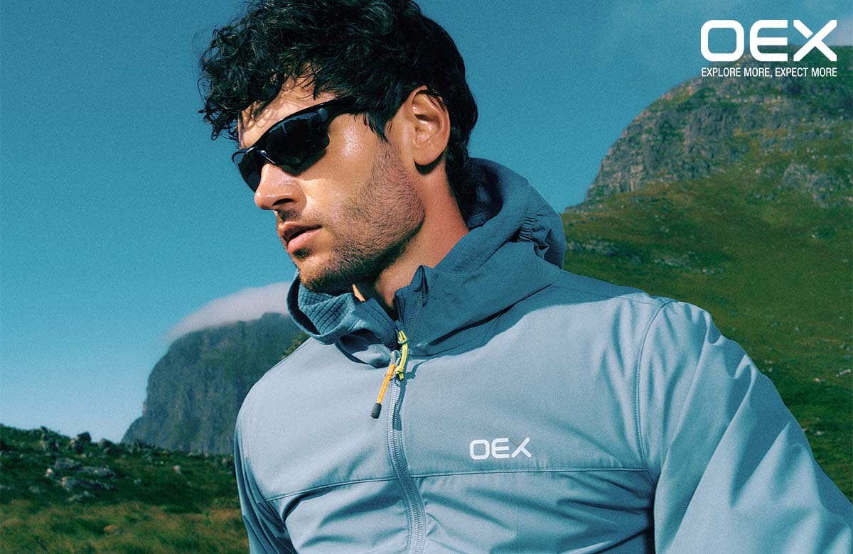 Man wearing OEX jacket and sunglasses with mountains in the background