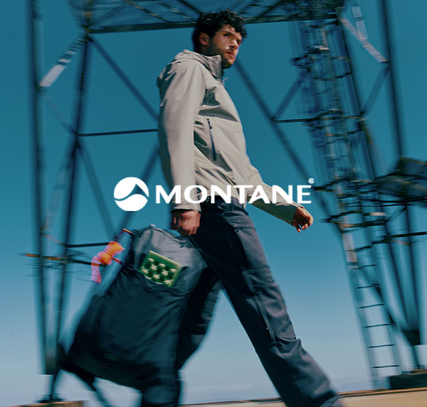 A person walks outdoors past a metal structure carrying a large duffel bag, wearing a light jacket, with the brand name “Montane” displayed across the image.