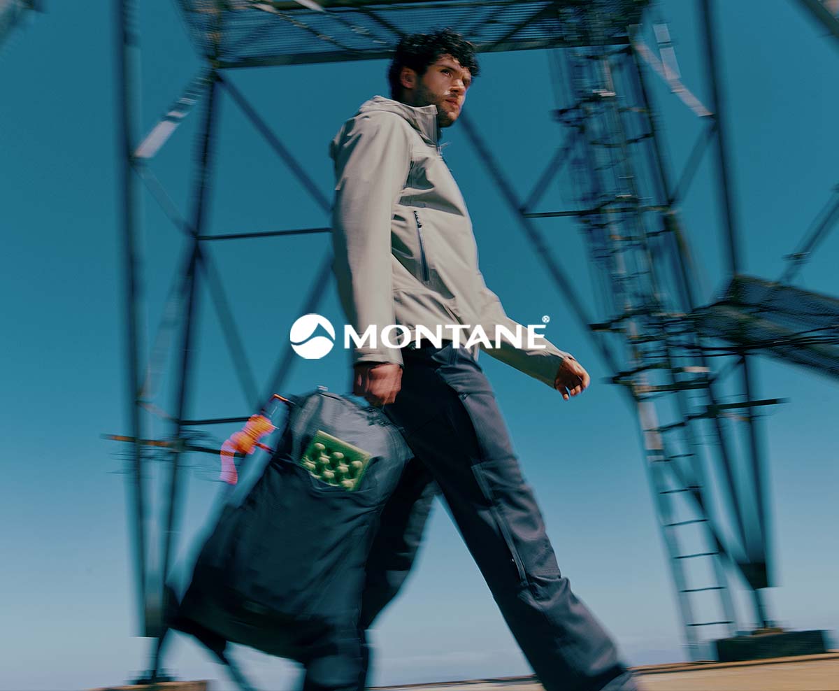 A person walks outdoors past a metal structure carrying a large duffel bag, wearing a light jacket, with the brand name “Montane” displayed across the image.