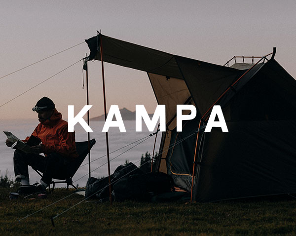 A campsite scene at dusk shows a tent set up on open ground, with a person seated nearby, and the brand name “Kampa” displayed prominently across the centre.
