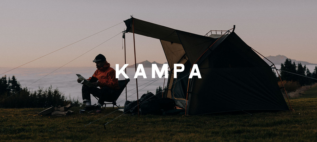 A campsite scene at dusk shows a tent set up on open ground, with a person seated nearby, and the brand name “Kampa” displayed prominently across the centre.