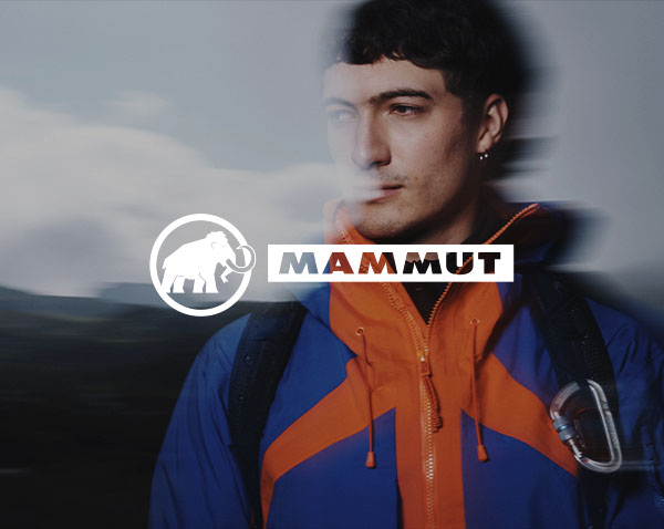 A person wearing a bright outdoor jacket and backpack stands in a blurred mountainous landscape, with the brand name “Mammut” displayed across the centre.