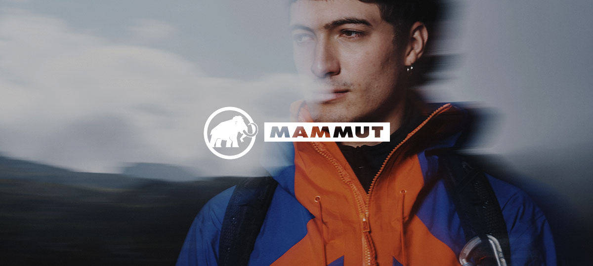 A person wearing a bright outdoor jacket and backpack stands in a blurred mountainous landscape, with the brand name “Mammut” displayed across the centre.