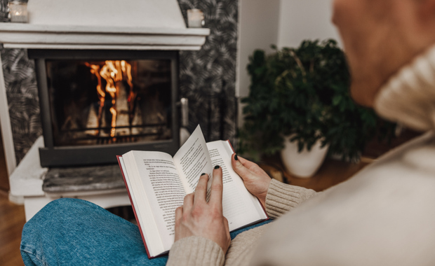 Henkilö lukee kirjaa nojatuolissa takkatulen ääressä.