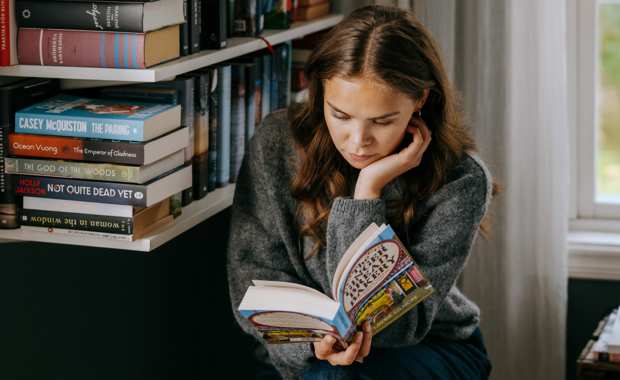 Nuori nainen istuu kirjahyllyn vieressä ja uppoutuu värikkääseen kirjaan rauhallisessa, luonnonvalossa olevassa huoneessa.