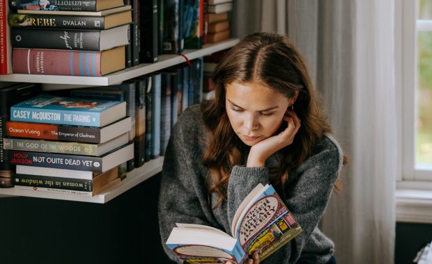 Kvinne med mørkt hår sitter foran en velfyllt bokhylle og leser i en pocketbok.