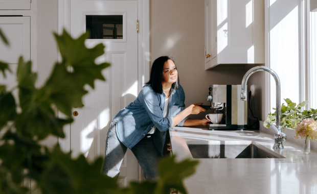 Nainen valmistaa kahvia valoisassa keittiössä, auringonvalo paistaa ikkunasta sisään ja kodikas tunnelma täyttää huoneen.
