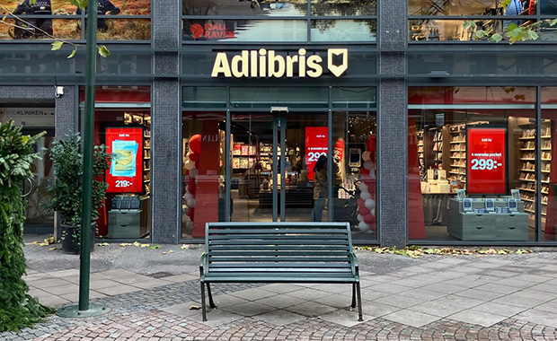 Adlibris bokhandel sedd utifrån, med upplyst entré, stora skyltfönster och bokhyllor och kampanjskyltar synliga inne i butiken.