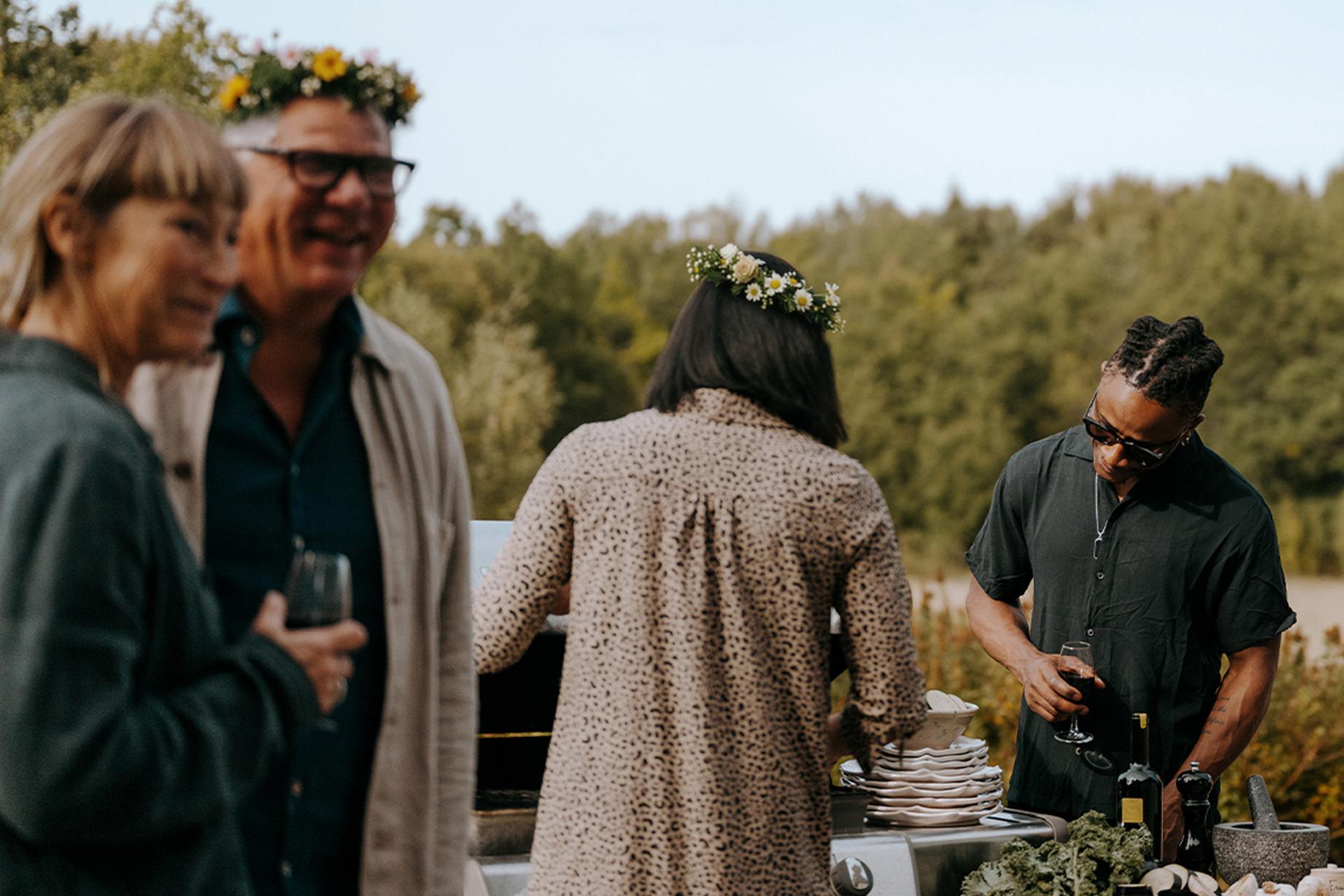 Bild av fyra personer utomhus vid en grill och ett matbord, där en person lagar mat medan de andra håller glas. I förgrunden syns tallrikar, grönkål, flaskor och en mortel, med skog i bakgrunden.
