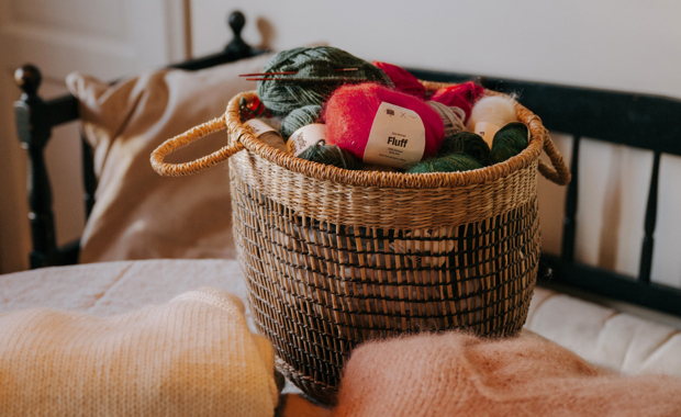 Korg fylld med garnnystan och stickor står på ett bord, omgiven av mjuka stickade tröjor i ett ombonat rum.