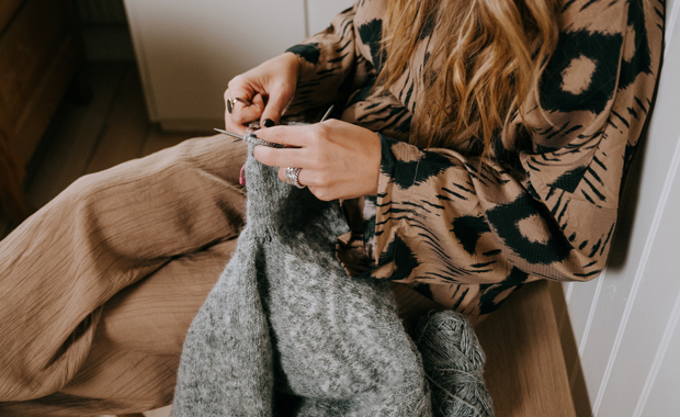 Person som sitter och stickar med grått garn, med pågående stickning i knät och ett garnnystan bredvid.