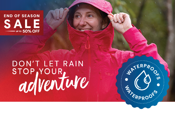 DON'T LET RAIN STOP YOUR ADVENTURE. A smiling woman pulls up the hood of a Rab Women's Downpour Mountain Jacket in Pink. 