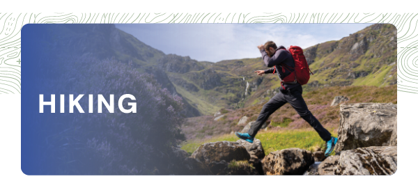 HIKING. A man jumps from a rock while hiking in a glen. He wears a black waterproof jacket and trousers, and blue walking shoes. 