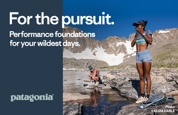 PATAGONIA: FOR THE PURSUIT. Performance Foundations For Your Wildest Days. Two women by a creek, mountains stretching in the background. In the foreground, a woman wearing Patagonia Trailblazer Shorts and a Patagonia Capilene Cool Daily Graphic T-Shirt uses her shirt to wipe sweat from her face. 