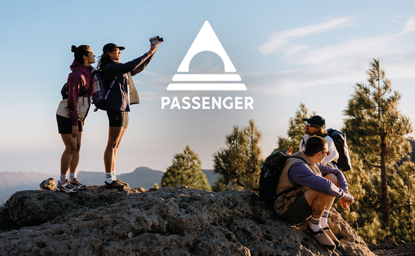 PASSENGER. Four people wearing Passenger clothing at the top of a hill in the sun, treetops visible in the background. On the left, two women take a selfie using a polaroid camera. On the right, two men sit on a rocky outcrop. One man looks out over the scenery, the other back at the two women. 