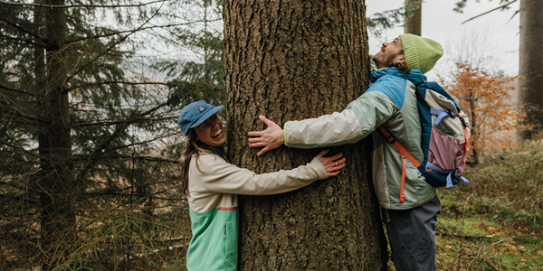 A man and a woman hug the trunk of a tree, laughing. Both are dressed in Passenger clothing.
