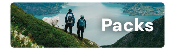 PACKS. A man and woman stand with a dog on a mountain overlooking a lake. Both wear Rab packs. 