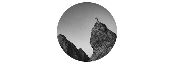 A black and white image of a climber standing at the peak of a rock, arms outstretched. 