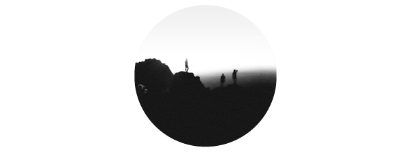 A black and white image of three climbers on a ridge, silhouetted against a foggy sky. 