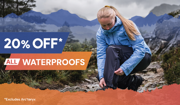 20% OFF ALL WATERPROOFS. A woman wearing a Rab Women's Downpour Mountain Jacket in Blue zips up the leg of her Rab Women's Downpour Pants.