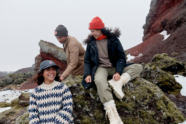 Drie personen dragen warme outdoor kleding op een rotsachtig berglandschap met sneeuwresten op de achtergrond.